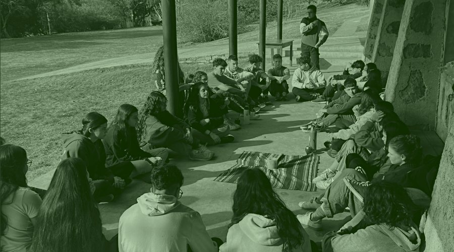 Una ronda de animadores sentados en el porche del salón multiusos durante un momento de oración en un
                                campamento de Animadores en el Campanero.